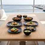 Ahşap tekne masasında deniz tarağı, ızgara ahtapot, caprese salatası ve enginar ikramlarıyla sunulan mat siyah döküm taş efektli kare kırılmaz melamin 8 parça Obsidian (M501208) 2 kişilik yemek takımı seti.