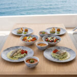 Ahşap tekne masasında ızgara balık, burrata salatası, çorba ve tatlı ikramlarıyla sunulan açık gri-mavi bulut mermer dokulu kırılmaz melamin 8 parça Velora Cloud (M506108) 2 kişilik yemek takımı seti.