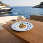 Ahşap tekne masasında zeytinyağı gezdirmesi, taze mikro filizler ve sarı yenilebilir çiçekler eşliğinde dilimlenmiş mühürlenmiş ton balığı tataki sunumuyla gösterilen mavi yaprak desenli açık gri zeminli kırılmaz melamin 6'lı yuvarlak tatlı, meze ve pasta tabağı seti (Ø 17,5 cm).