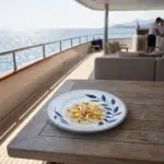 Lüks yatın ahşap güverte masasında ince zeytinyağı sosu, narenciye/limon dilimleri ve taze mikro otlar eşliğinde sunulan levrek carpaccio ikramıyla gösterilen mavi yaprak desenli açık gri zeminli kırılmaz melamin 3'lü büyük boy yuvarlak servis ve ana yemek tabağı seti (Ø 25 cm).