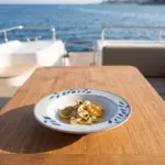 Gün batımı ışığında açık denizde seyir halindeki lüks yatın ahşap güverte masasında ızgara kalamar dilimleri, ızgara limon dilimleri, taze kekik ve sızma zeytinyağı sosu eşliğinde sunulan mavi yaprak desenli açık gri zeminli kırılmaz melamin 3'lü yuvarlak çukur yemek ve çorba tabağı seti (Ø 20,3 cm).