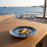 Gün batımı ışığında açık denizde seyreden lüks yatın ahşap güverte masasında tavada mühürlenmiş beyaz etli balık filetosu, iki adet mühürlenmiş deniz tarağı (scallop), taze biberiye ve otlu zeytinyağı gezdirmesi eşliğinde sunulan açık krem çerçeveli reaktif okyanus mavisi efektli kırılmaz melamin 3'lü yuvarlak yemek ve servis tabağı seti (Ø 23 cm).