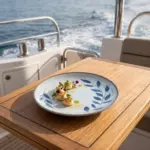 Seyir halindeki lüks yatın ahşap güverte masasında mühürlenmiş deniz tarağı (scallop) ikramı, taze otlar, sızma zeytinyağı ve mor-beyaz yenilebilir çiçekler eşliğinde gösterilen mavi yaprak desenli açık gri zeminli kırılmaz melamin 3'lü yuvarlak yemek ve servis tabağı seti (Ø 23 cm).