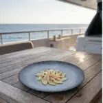 Seyir halindeki lüks yatın ahşap güverte masasında taze levrek carpaccio/sashimi dilimleri, sızma zeytinyağı, limon kabuğu rendesi ve ince kıyılmış taze otlar eşliğinde sunulan dairesel spral dokulu lacivert-mavi mermer efektli kırılmaz melamin 3'lü yuvarlak büyük boy servis ve düz ana yemek tabağı seti (Ø 25 cm).