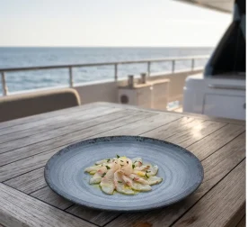 Seyir halindeki lüks yatın ahşap güverte masasında taze levrek carpaccio/sashimi dilimleri, sızma zeytinyağı, limon kabuğu rendesi ve ince kıyılmış taze otlar eşliğinde sunulan dairesel spral dokulu lacivert-mavi mermer efektli kırılmaz melamin 3'lü yuvarlak büyük boy servis ve düz ana yemek tabağı seti (Ø 25 cm).