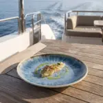 Seyir halindeki lüks yatın ahşap güverte masasında taze marine edilmiş levrek/akya sashimi dilimleri, taze kekik yaprakları ve sızma zeytinyağı sosu eşliğinde sunulan açık krem çerçeveli reaktif okyanus mavisi efektli kırılmaz melamin 3'lü yuvarlak büyük boy servis ve düz ana yemek tabağı seti (Ø 25 cm).