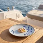 Açık denizde seyir halindeki lüks yatın ahşap güverte masasında taze deniz tarağı (scallop) crudo dilimleri ve renkli yenilebilir çiçekler eşliğinde sunulan mavi yaprak desenli açık gri zeminli kırılmaz melamin 3'lü yuvarlak büyük boy servis ve düz ana yemek tabağı seti (Ø 25 cm).