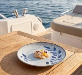 Açık denizde seyir halindeki lüks yatın ahşap güverte masasında taze deniz tarağı (scallop) crudo dilimleri ve renkli yenilebilir çiçekler eşliğinde sunulan mavi yaprak desenli açık gri zeminli kırılmaz melamin 3'lü yuvarlak büyük boy servis ve düz ana yemek tabağı seti (Ø 25 cm).