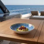 Ahşap tekne masasında patates/fava püreresi yatağında zeytinyağı sosu, küp patatesler ve pembe-mor yenilebilir çiçekler eşliğinde sunulan ızgara ahtapot ikramıyla gösterilen açık krem çerçeveli reaktif okyanus mavisi efektli kırılmaz melamin 3'lü yuvarlak geniş çukur yemek, makarna ve çorba tabağı seti (Ø 23 cm, 540 ml).
