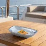 Gün batımı ışığında açık denizde seyir halindeki lüks yatın ahşap güverte masasında ızgara limon dilimi, taze kekik ve zeytinyağı eşliğinde sunulan ızgara karides ikramı ile gösterilen mavi yaprak desenli açık gri zeminli kırılmaz melamin 6'lı küçük boy kayık meze ve zeytinyağlı tabağı seti (14x8,5 cm).