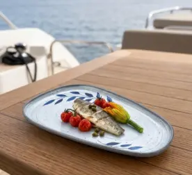 Ahşap tekne masasında ızgara balık filetosu, fırınlanmış salkım çeri domatesler, kabak çiçeği dolması ve kapari taneleri eşliğinde sunulan mavi yaprak desenli açık gri zeminli kırılmaz melamin 3'lü asimetrik kayık kayık servis ve meze tabağı seti (27x15 cm).