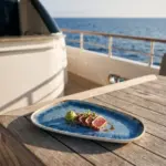 Seyir halindeki lüks yatın ahşap güverte masasında susamlı ton balığı tataki dilimleri, vasabi/avokado püre küresi, soyalı glaze sosu ve taze mikro filizler eşliğinde gösterilen açık krem çerçeveli reaktif okyanus mavisi efektli kırılmaz melamin 3'lü büyük boy asimetrik kayık sunum ve servis tabağı seti (33x18 cm).