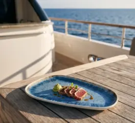 Seyir halindeki lüks yatın ahşap güverte masasında susamlı ton balığı tataki dilimleri, vasabi/avokado püre küresi, soyalı glaze sosu ve taze mikro filizler eşliğinde gösterilen açık krem çerçeveli reaktif okyanus mavisi efektli kırılmaz melamin 3'lü büyük boy asimetrik kayık sunum ve servis tabağı seti (33x18 cm).