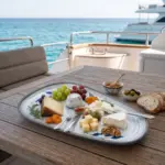 Turkuaz deniz manzaralı lüks yatın ahşap güverte masasında Brie, Manchego, keçi peyniri çeşitleri, yeşil ve siyah üzüm, taze incir, kuru kayısı, ceviz, biberiye ve ekmek dilimleri eşliğinde zengin şarküteri/peynir tabağı sunumuyla gösterilen mavi yaprak desenli açık gri zeminli kırılmaz melamin 3'lü büyük boy asimetrik kayık sunum ve servis tabağı seti (33x18 cm).