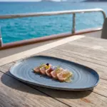Turkuaz deniz manzaralı lüks yatın ahşap güverte masasında mühürlenmiş ton balığı tataki/sashimi dilimleri, yeşil otlu jel damlaları, taze dereotu ve mor-sarı yenilebilir çiçekler eşliğinde gösterilen dairesel spral dokulu lacivert-mavi mermer efektli kırılmaz melamin 2'li ekstra büyük boy (battal) asimetrik kayık servis ve sunum tabağı seti (37,5x20 cm).