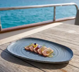 Turkuaz deniz manzaralı lüks yatın ahşap güverte masasında mühürlenmiş ton balığı tataki/sashimi dilimleri, yeşil otlu jel damlaları, taze dereotu ve mor-sarı yenilebilir çiçekler eşliğinde gösterilen dairesel spral dokulu lacivert-mavi mermer efektli kırılmaz melamin 2'li ekstra büyük boy (battal) asimetrik kayık servis ve sunum tabağı seti (37,5x20 cm).