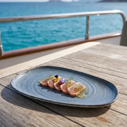 Turkuaz deniz manzaralı lüks yatın ahşap güverte masasında mühürlenmiş ton balığı tataki/sashimi dilimleri, yeşil otlu jel damlaları, taze dereotu ve mor-sarı yenilebilir çiçekler eşliğinde gösterilen dairesel spral dokulu lacivert-mavi mermer efektli kırılmaz melamin 2'li ekstra büyük boy (battal) asimetrik kayık servis ve sunum tabağı seti (37,5x20 cm).