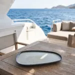 Derin mavi deniz ve ada manzaralı lüks yatın ahşap güverte masasında gösterilen koyu kenar çerçeveli buz mavisi ve gri çatlatmalı sır (crackle glaze) efektli, ekstra büyük boy (battal) kırılmaz melamin 2'li asimetrik kayık servis, balık ve meze tabağı seti (37,5x20 cm).