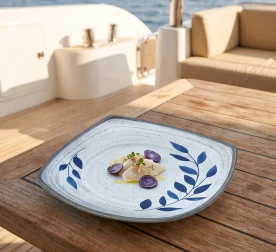 Gün batımı ışığında açık denizde seyir halindeki lüks yatın ahşap güverte masasında taze levrek crudo/sashimi dilimleri, mor patates/turp dilimleri, taze mikro filizler ve sızma zeytinyağı eşliğinde sunulan mavi yaprak desenli açık gri zeminli kırılmaz melamin 3'lü kare büyük boy servis ve ana yemek tabağı seti (24,1x24,1 cm).