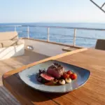 Gün batımı ışığında açık denizde seyir halindeki lüks yatın ahşap güverte masasında demi-glace sos yatağında orta-az pişmiş dana biftek/bonfile dilimleri, fırınlanmış salkım çeri domatesler, karamelize arpacık soğanlar ve mor taze filizler eşliğinde sunulan dairesel spral dokulu lacivert-mavi mermer efektli kırılmaz melamin 3'lü kare büyük boy servis ve ana yemek tabağı seti (26,4x26,4 cm).
