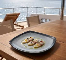 Gün batımı ışığında açık denizde seyir halindeki lüks yatın ahşap güverte masasında taze levrek carpaccio/sashimi dilimleri, taze kekik, sızma zeytinyağı ve renkli yenilebilir çiçek taç yaprakları eşliğinde gösterilen koyu kenarlı buz mavisi-gri çatlatmalı sır (crackle glaze) efektli kırılmaz melamin 3'lü kare büyük boy servis tabağı seti (26,4x26,4 cm).