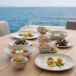 Ahşap tekne masasında deniz tarağı, burrata salatası, çorba, karides ve ikramlıklarla sunulan otantik altın-bronz eskitme desenli kare kırılmaz melamin 8 parça Old Ivory (M503208) 2 kişilik yemek takımı seti.