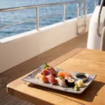Ahşap tekne masasında zencefil turşusu, wasabi ve soya sosu eşliğinde sashimi, nigiri ve maki sushi tabağı sunumuyla gösterilen açık gri-mavi bulut mermer dokulu büyük boy kırılmaz melamin Velora Cloud 3'lü dikdörtgen servis ve sunum tabağı seti (30 x 20,7 cm).