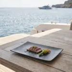 Ahşap tekne masasında mühürlenmiş ton balığı tataki, kabak çiçeği dolması ve fesleğen yağı sunumuyla gösterilen koyu toprak tonlarında mat kil dokulu büyük boy kırılmaz melamin Velora Clay 3'lü dikdörtgen servis ve sunum tabağı seti (30 x 20,7 cm).