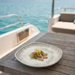 Ahşap tekne masasında limon dilimleri ve taze maydanoz eşliğinde marine balık carpaccio sunumuyla gösterilen otantik altın-bronz eskitme desenli kare/yuvarlak yumuşak hatlı kırılmaz melamin Old Ivory 6'lı pasta ve tatlı tabağı seti (18 x 18 cm).