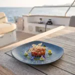 Ahşap tekne masasında avokado püresi, çıtır kıtır dantecelle ve yenilebilir çiçekler eşliğinde ton balığı tartar (tuna tartar) sunumuyla gösterilen dumanlı mavi taş efektli kare/yuvarlak yumuşak hatlı kırılmaz melamin Velora Mist 3'lü servis ve yemek tabağı seti (22 x 22 cm).
