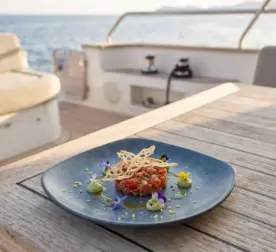 Ahşap tekne masasında avokado püresi, çıtır kıtır dantecelle ve yenilebilir çiçekler eşliğinde ton balığı tartar (tuna tartar) sunumuyla gösterilen dumanlı mavi taş efektli kare/yuvarlak yumuşak hatlı kırılmaz melamin Velora Mist 3'lü servis ve yemek tabağı seti (22 x 22 cm).